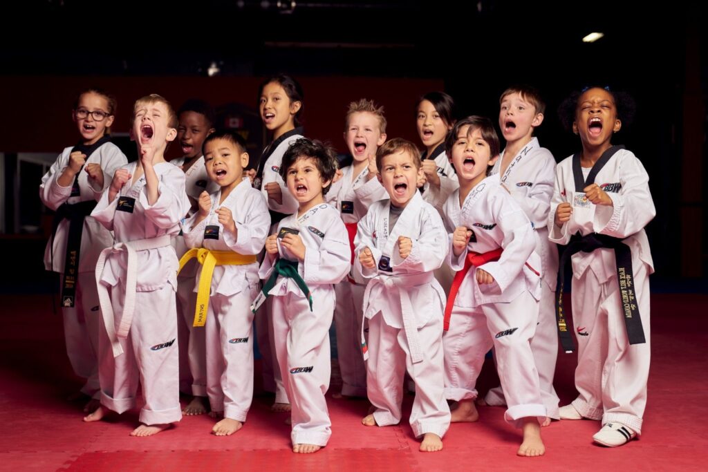 A diverse group of young martial arts students wearing white uniforms and various colored belts practicing their kihap shouts during a kids' karate class.