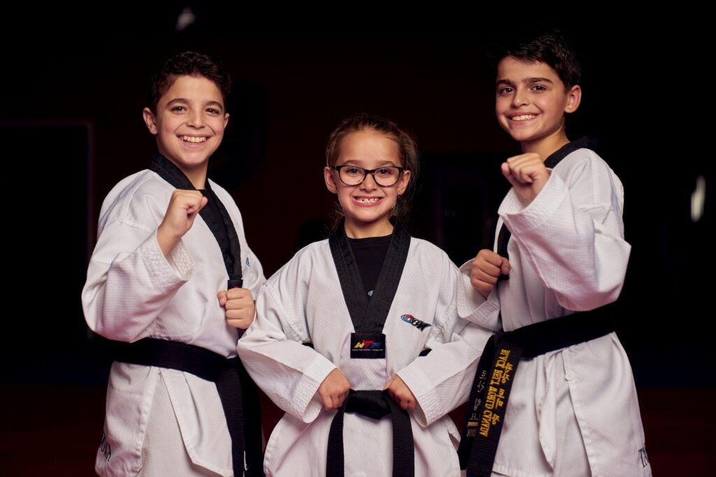 Three smiling young students in white uniforms earning their martial arts black belt while practicing youth self-defense techniques.