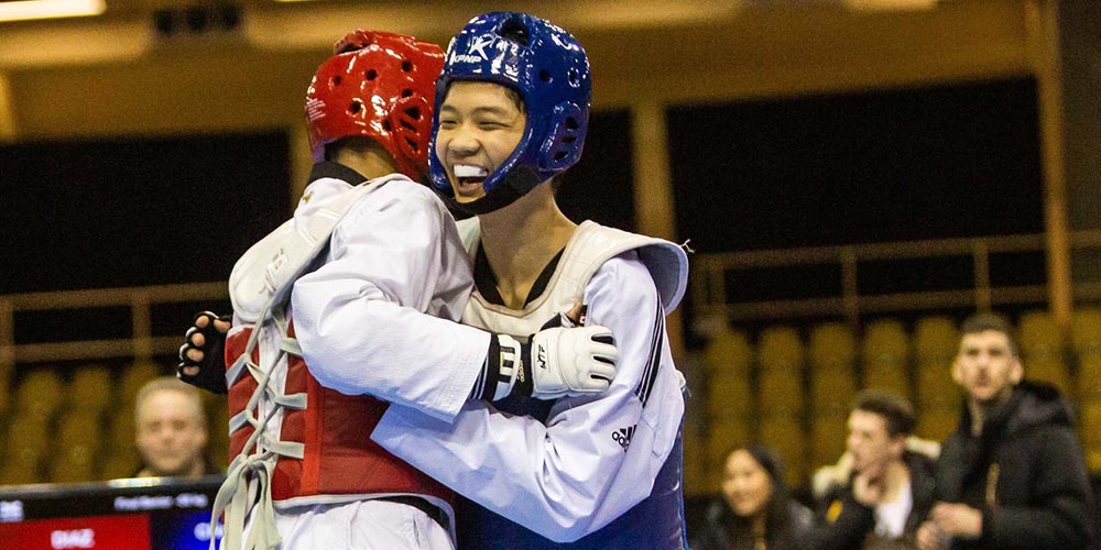 Two young competitors in protective gear showing sportsmanship in martial arts through a hug after a youth Taekwondo tournament.